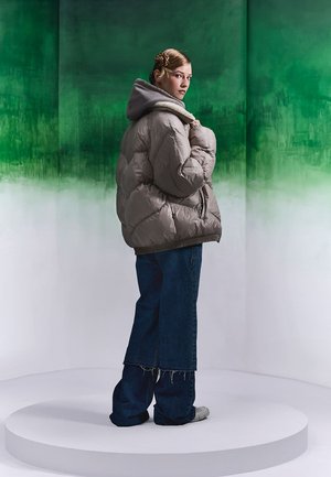 Overmaat, gewatteerde grijze pufferjack met capuchon, gecombineerd met wijde, versleten spijkerbroek en grijze sneakers, tegen een groene achtergrond.