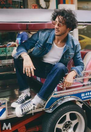 Jeansjacke mit sichtbaren Nähten, weißes Tanktop, dunkle Jeans und schwarze Sneakers mit weißen Sohlen, sitzend in einem bunten Tuk-Tuk.