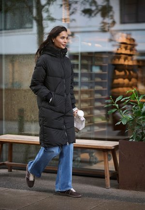 Lång svart vadderad jacka med dragkedja framtill, huva och sidofickor; kombinerad med vida blå jeans och mörkbruna skor.