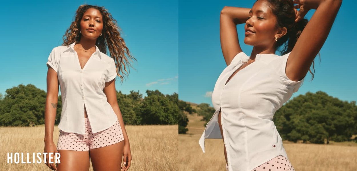Junge Frau trägt eine weiße kurzärmelige Bluse und rosa gepunktete Shorts, posiert draußen auf einem sonnigen Feld mit Bäumen im Hintergrund.