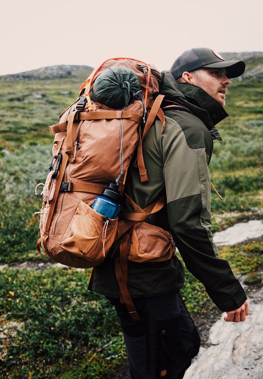 Brun vandringsryggsäck med flera fickor, en hållare för vattenflaska på sidan och en grön sovsäck fäst; tillverkad av hållbart och väderbeständigt material.