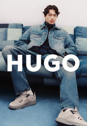 Chaqueta de mezclilla y jeans en azul claro, combinados con zapatillas deportivas beige. El modelo está sentado en un sofá multicolor. Texto en negrita "HUGO" abajo.