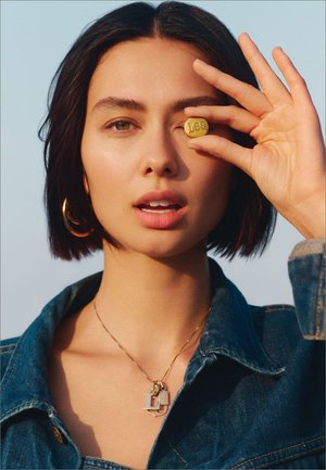Accessorio simile a una moneta d'oro con "Leo" inciso, tenuto contro l'occhio di una persona. Indossa una giacca di jeans e collane sovrapposte. Tono di pelle naturale.