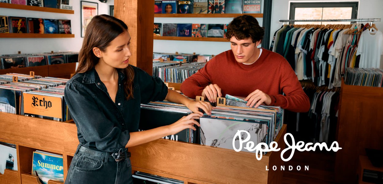 Zwei Personen durchstöbern Schallplatten in einer hölzernen Vitrine. Die Frau trägt ein dunkles Jeanshemd, und der Mann trägt einen roten Pullover. Die Schallplatten sind nach Labels sortiert.