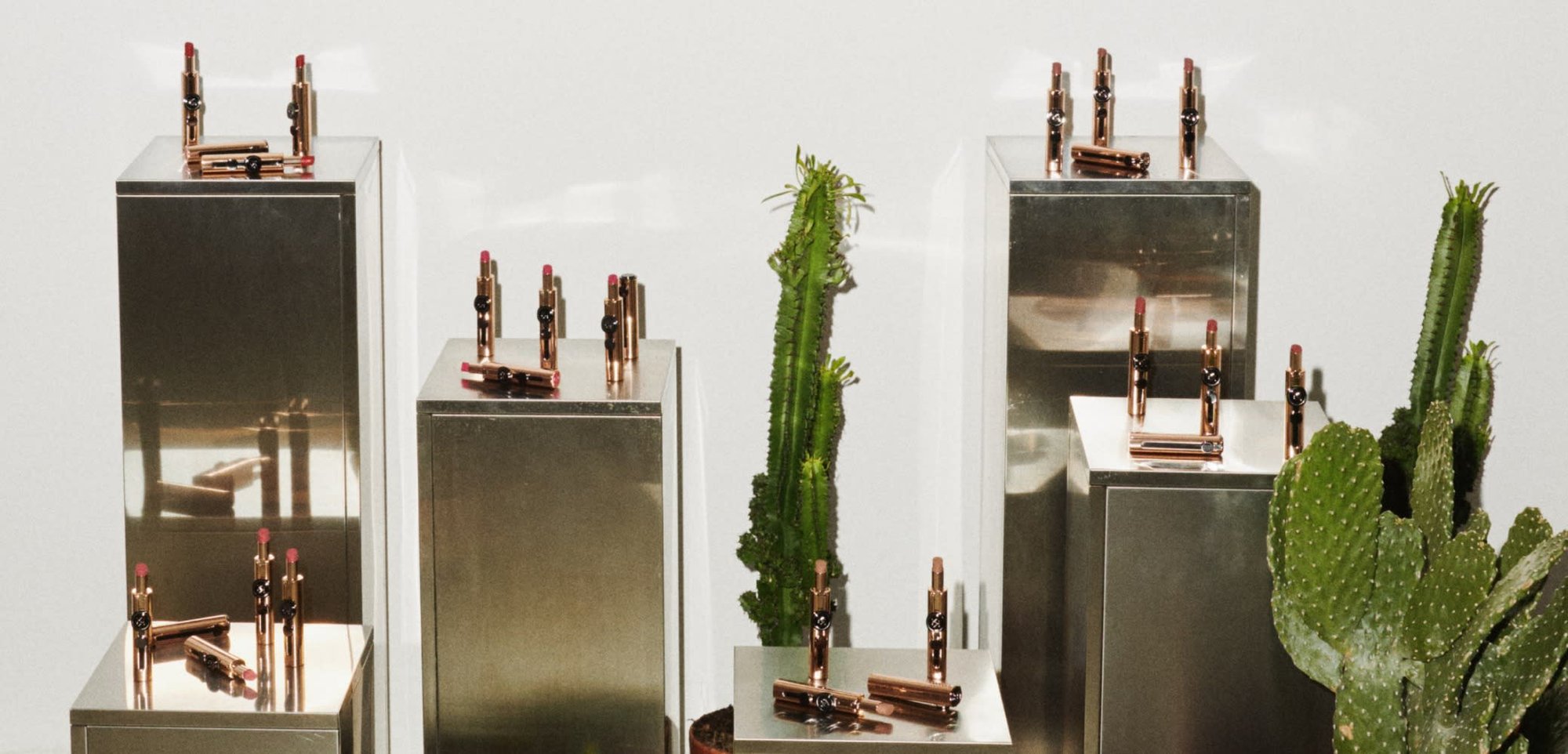 Eight lipstick tubes with rose gold accents arranged on metallic display pedestals, accompanied by cacti and small potted plants on a sandy surface.
