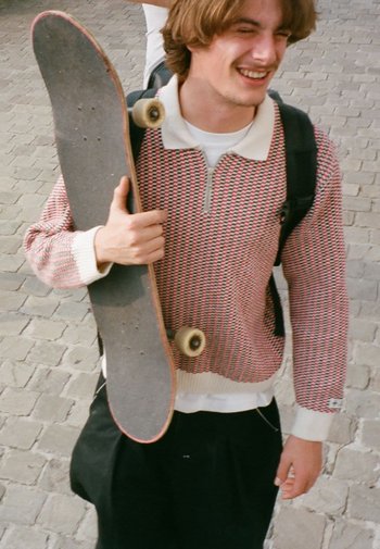 Gebreide trui met rood en wit geometrisch patroon, rits, beige kraag; vasthoudend aan een grijze skateboard met zichtbare wielen. Casual zwarte broek.