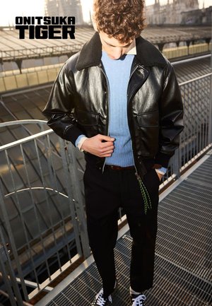 Giacca bomber in pelle nera con colletto morbido, abbinata a un maglione azzurro chiaro e pantaloni neri; presenta dettagli con cerniera verde e sneakers.