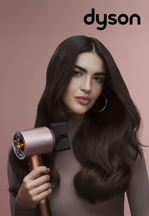 Femme aux longs cheveux bruns tenant un sèche-cheveux Dyson près de cheveux volumineux coiffés sur un fond rose avec le logo Dyson.