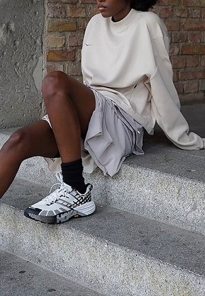 Weiße Hoka-Sneaker mit schwarz-weißem kariertem Muster und gelben Akzenten, kombiniert mit einem lockeren beigen Sweatshirt und einem grauen, geschichteten Rock.