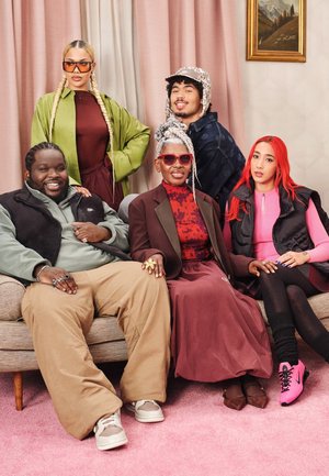 Cinque persone sedute su un divano beige. Indossano abiti di vari colori, tra cui verde, rosa e bordeaux. Uno sfondo rosa.