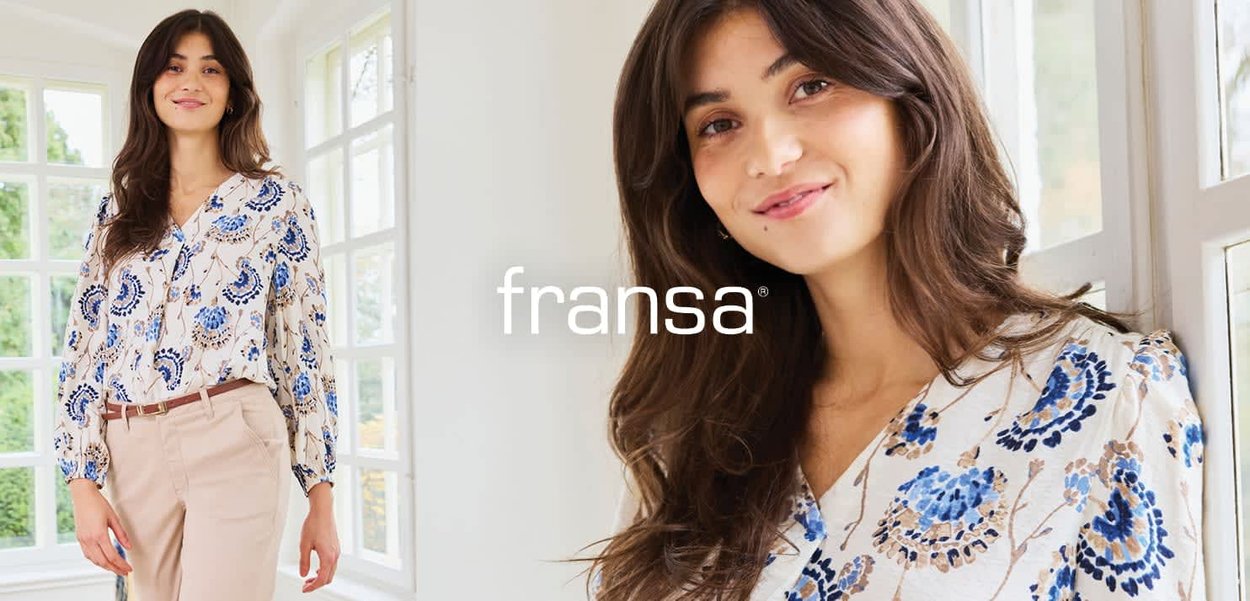 Junge Frau mit langen braunen Haaren trägt eine geblümte blaue und beige Bluse sowie beige Hosen in einem hellen, von Fenstern durchfluteten Raum.