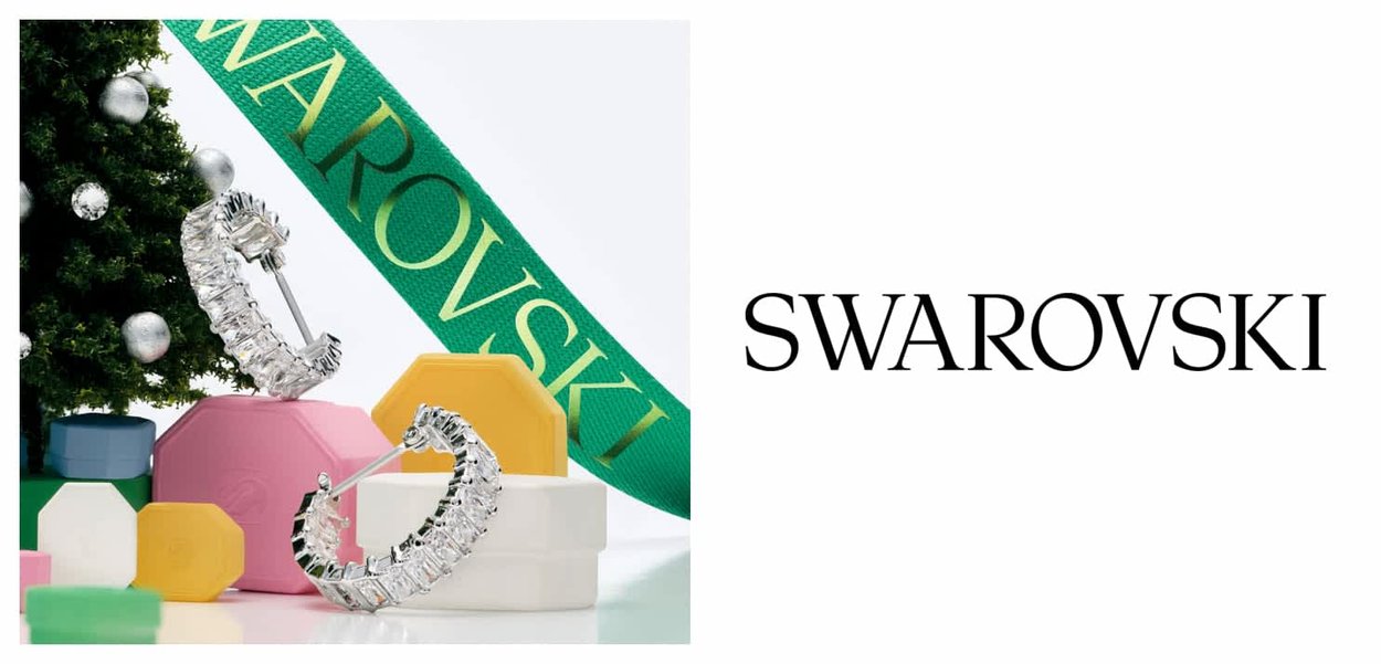 Des boucles d'oreilles en cristal Swarovski sont exposées parmi des boîtes géométriques colorées et un sapin de Noël, avec un ruban vert portant le nom de la marque.