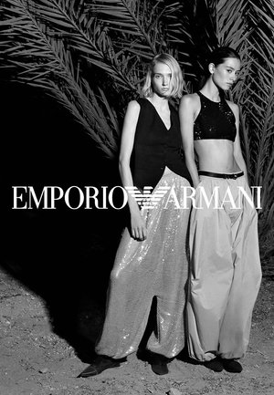 Dos modelos femeninas están junto a grandes hojas de palma, vistiendo tops sin mangas de alta moda y pantalones anchos en un ambiente en blanco y negro de Emporio Armani.