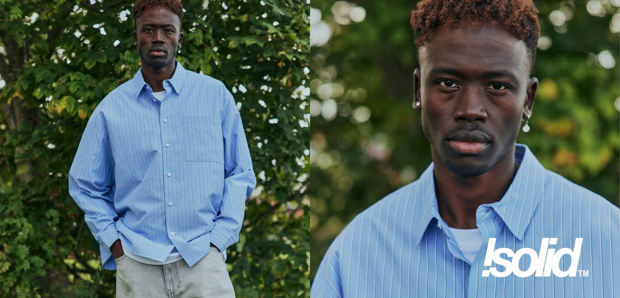 Junger Mann mit kurzen lockigen Haaren, der ein blau gestreiftes Hemd und helle Hosen trägt, steht im Freien vor einem grünen, blättrigen Hintergrund.