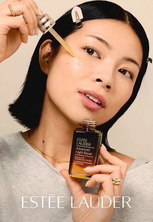 Kvinde påfører Estée Lauder Advanced Night Repair serum på kinden med en dråbetæller og holder flasken tæt ved ansigtet.