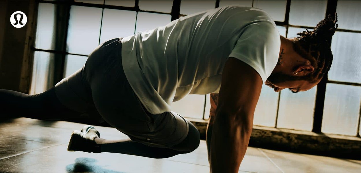 Mężczyzna w sportowym stroju wykonujący ćwiczenie górski wspinacz w pomieszczeniu przy dużych oknach, ubrany w białą koszulkę i ciemne legginsy.