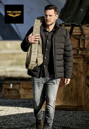 Chaqueta acolchada negra sobre una camisa oscura, vaqueros claros, llevando una bolsa de lona beige. Telón de fondo industrial con cajas de madera y estructuras metálicas.