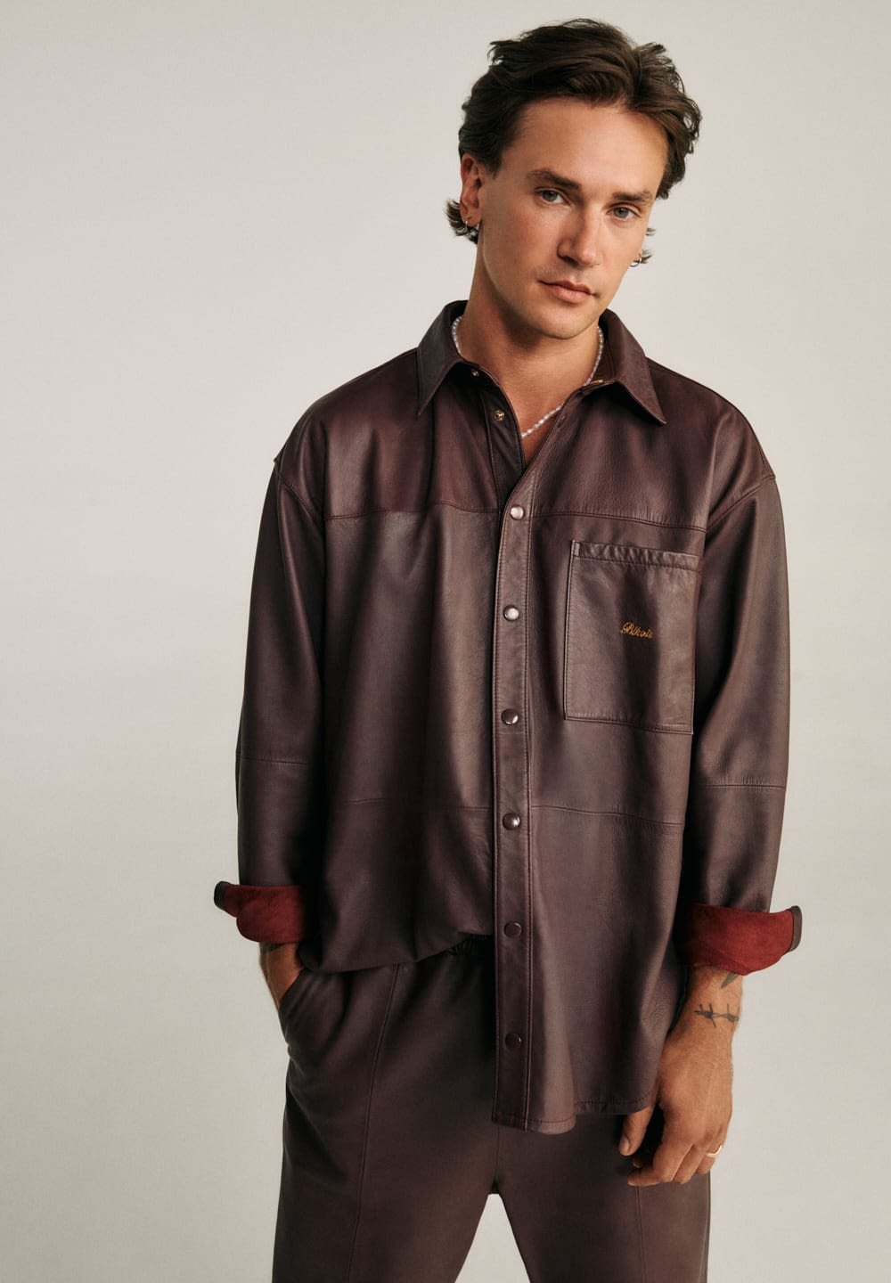 Młody mężczyzna ubrany w ciemnobrązową skórzaną koszulę i spodnie, z podwiniętymi rękawami i rękami w kieszeniach, stojący na jednolitym tle.