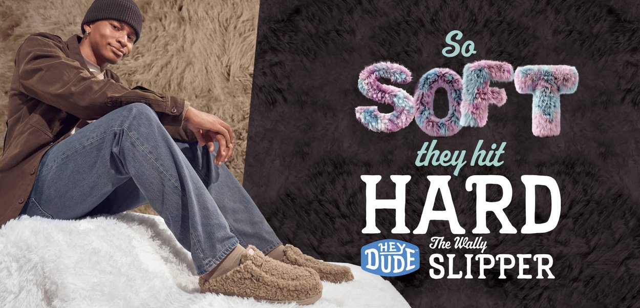 Pantofole beige, testurizzate, con un esterno morbido e soffice e un piccolo dettaglio a bottone, indossate con jeans rilassati e una giacca marrone.