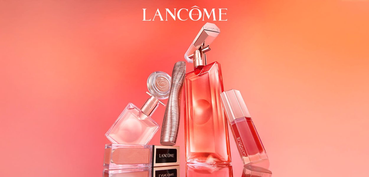 Bottiglie di profumo Lancome, mascara e lucidalabbra disposti su una superficie riflettente con uno sfondo sfumato rosa-arancio.
