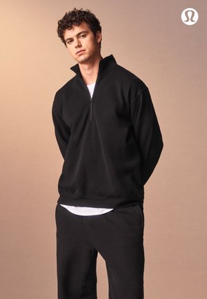 Zwarte quarter-zip sweater gemaakt van zacht, geborsteld materiaal met lange mouwen, gecombineerd met bijpassende zwarte joggers en een wit ondershirt.