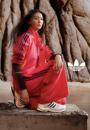 Młoda kobieta w jaskraworóżowym dresie Adidas i białych sneakersach pozuje siedząc na betonowym bloku na tle skalistego krajobrazu.