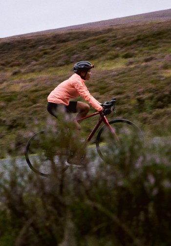 Ciclist pe o bicicletă roșie, purtând o jachetă roz aprins și pantaloni scurți negri, navigând pe un drum rural printre vegetația verde și violet.