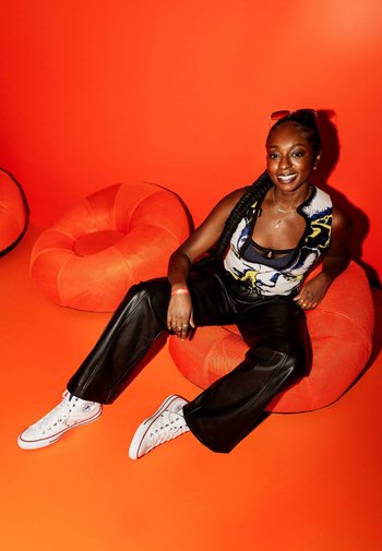 A patterned sleeveless top with black pants is worn with white sneakers. The subject sits on vibrant orange bean bags against an orange background.