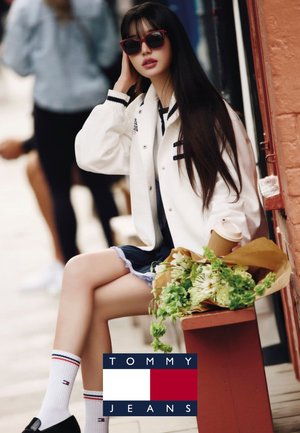 Młoda kobieta o długich ciemnych włosach, w okularach przeciwsłonecznych, białej kurtce i skarpetkach, siedząca na ławce, trzymająca filiżankę kawy i zielony bukiet kwiatów.