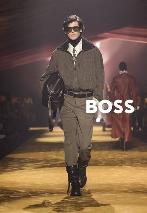 Modello maschile che cammina sulla passerella indossando una giacca marrone oversize con zip, cravatta stampata, pantaloni kaki, stivali neri, occhiali da sole, porta una grande borsa nera con logo BOSS.