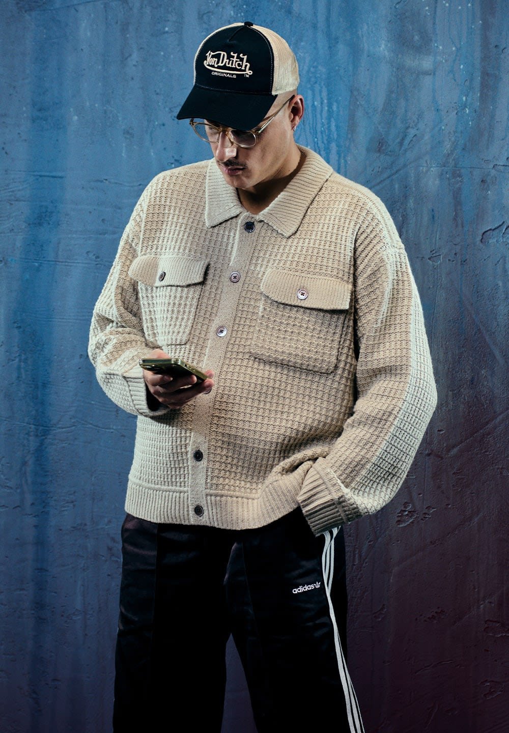 Šviesiai smėlinis tekstūruotas megztas švarkas su mygtukais priekyje, dviem krūtinės kišenėmis ir plačiais rankogaliais, derinamas su juodomis treningo kelnėmis ir kepure. Laiko išmanųjį telefoną.