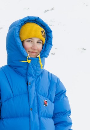 Helle blaue Daunenjacke mit hohem Kragen, ausgestattet mit gelben Kordeln und einem Logo-Patch, kombiniert mit einer gelben Strickmütze.