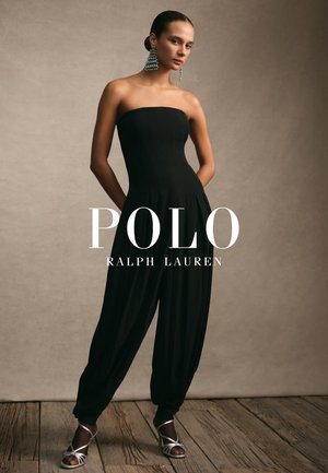 Combinaison noire sans bretelles avec un corsage ajusté et un pantalon drapé et ample. Le mannequin porte des talons élégants argentés et des boucles d'oreilles turquoise remarquables.