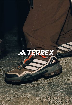 Chaussures de randonnée Terrex en brun et gris, avec une tige texturée, des rayures blanches, des accents orange et une semelle extérieure noire robuste.