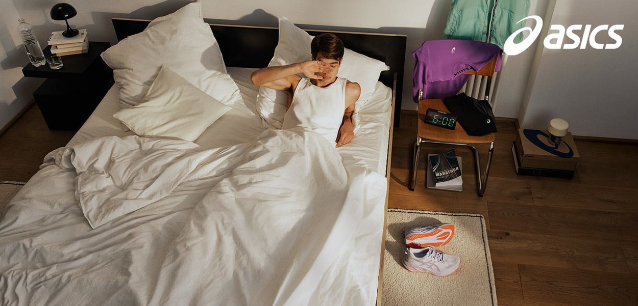 White bedding with a person in a tank top in bed. Nearby are running shoes, a clock, and clothing draped over a chair on a wooden floor.