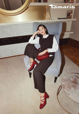 Rode hoge hakken met gekruiste bandjes, gecombineerd met donkere broek en een zwart vest over een wit shirt, zittend in een blauwe fauteuil.