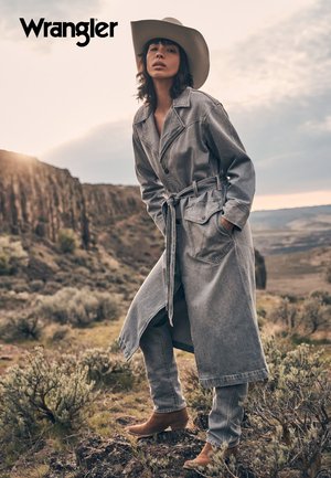 Cappotto trench in denim con vita cintata, abbinato a jeans a gamba larga coordinati e stivali alla caviglia marroni; sullo sfondo di un paesaggio rugged.