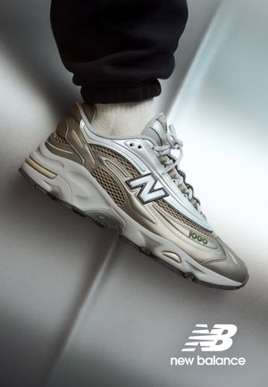 Sneaker beige clair New Balance en matériaux en maille et en cuir, avec un logo N proéminent et un détail vert 1000, semelle texturée.