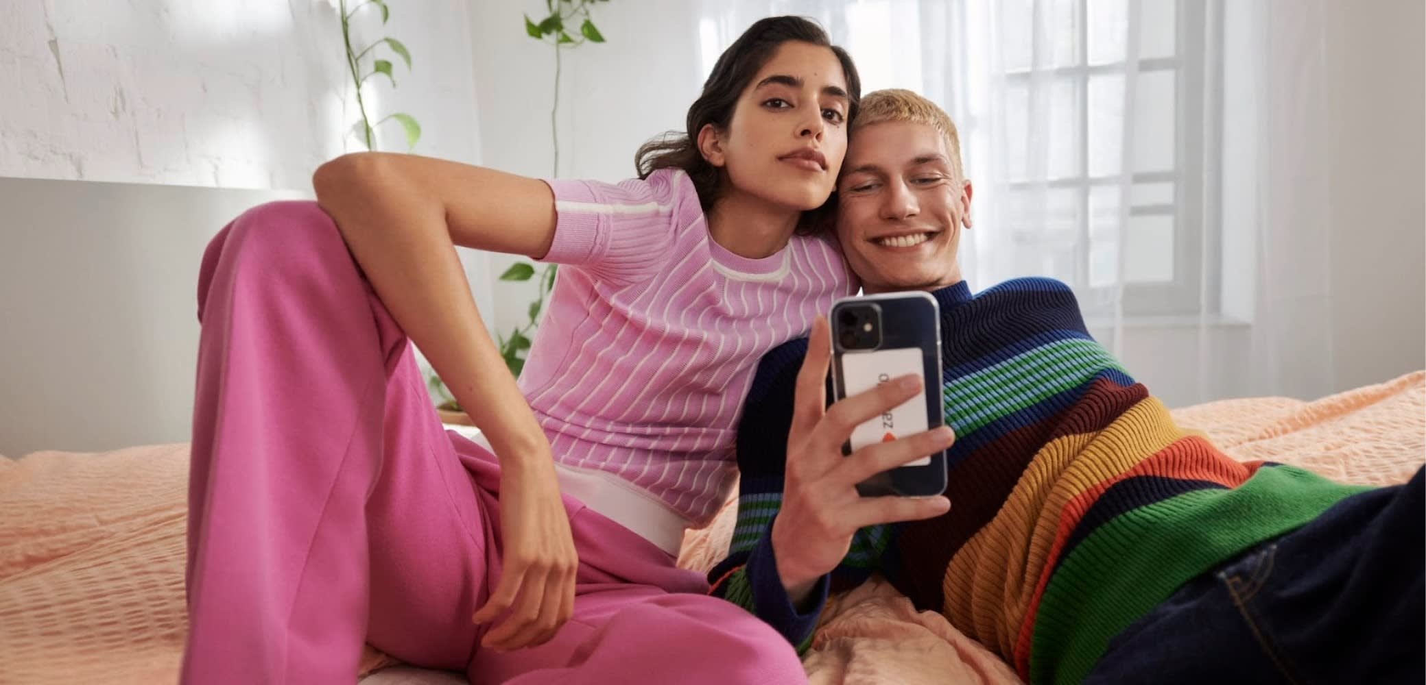 Coppia su un letto, che indossa un maglione a coste di un leggero rosa e pantaloni rosa, accanto a un maglione a righe blu scuro con accenti arcobaleno, mentre tiene un telefono.
