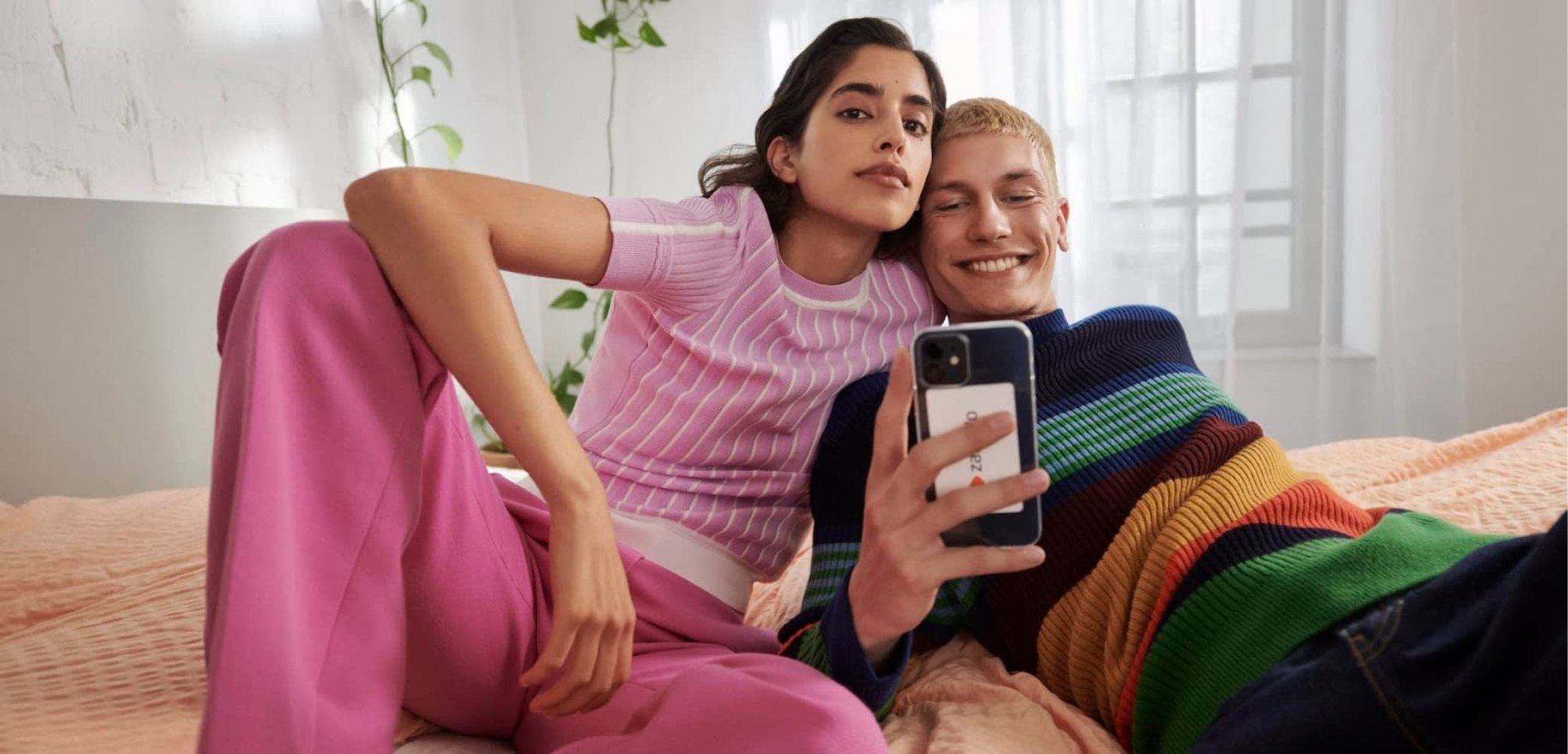 Deux personnes sont assises ensemble sur un lit. L'une porte un haut côtelé rose clair et un pantalon rose assorti ; l'autre est habillé d'un pull rayé multicolore.