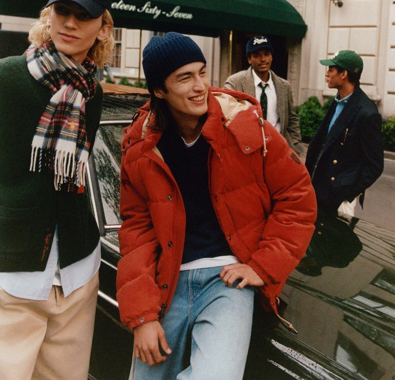 Veste matelassée rouge avec un col montant, portée sur un pull bleu marine. Fond urbain pittoresque avec une voiture sombre. Groupe de quatre personnes en tenue décontractée.