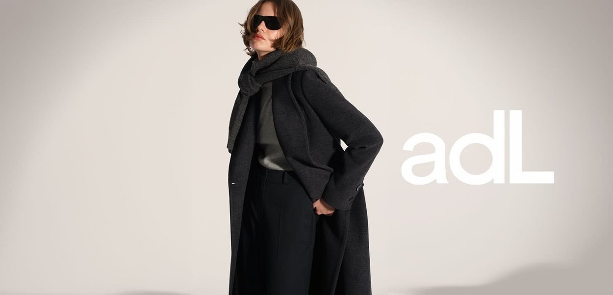 Vrouw met donkere zonnebril, zwarte lange jas, grijze sjaal en zwarte broek met hoge taille poseert tegen een lichte achtergrond met het "adL" logo.
