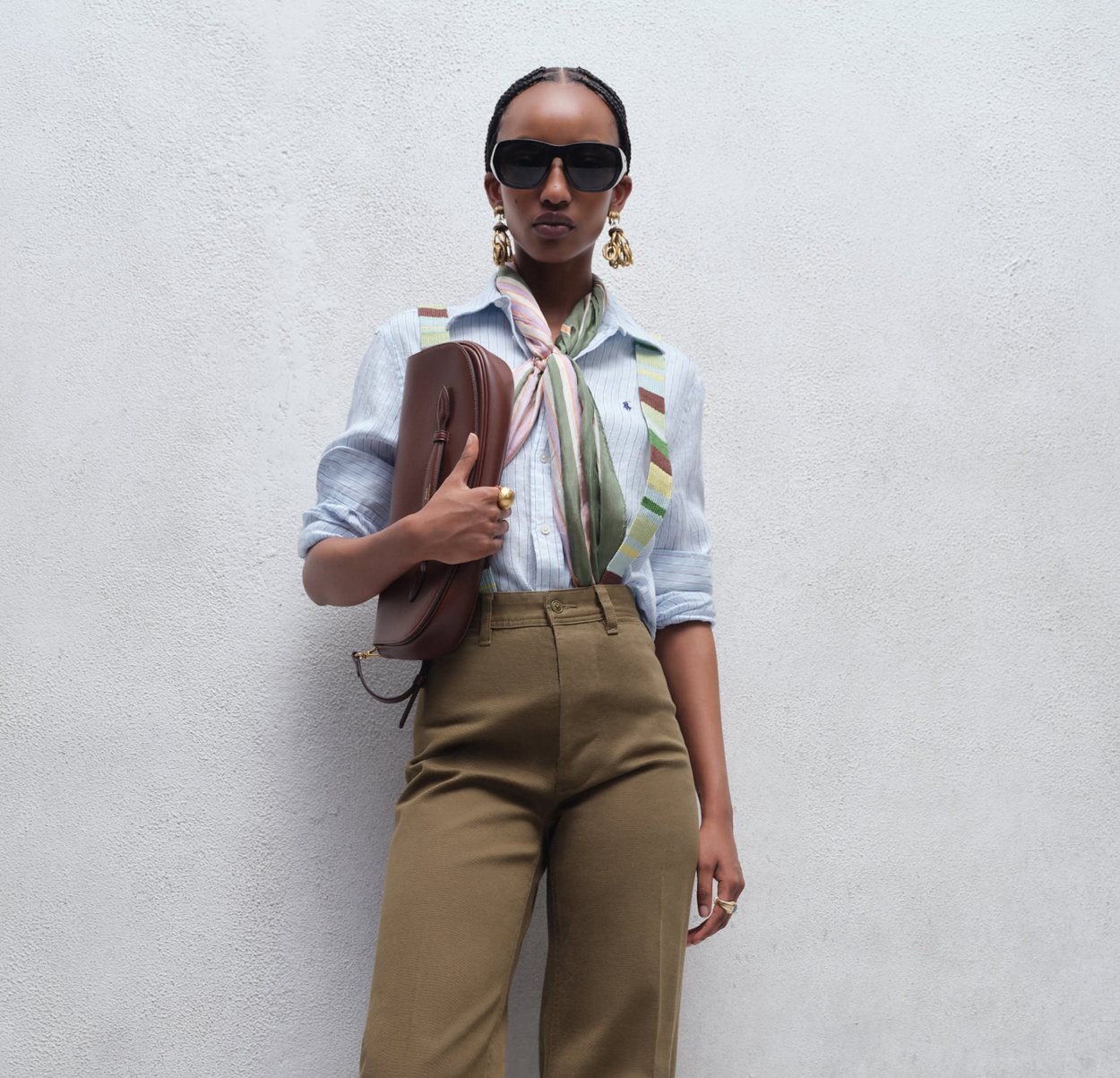 Kvinne med flette i håret som bærer solbriller, stripete skjorte, mønstrete skjerf, olivengrønne bukser, holder en brun clutch mot en hvit vegg.