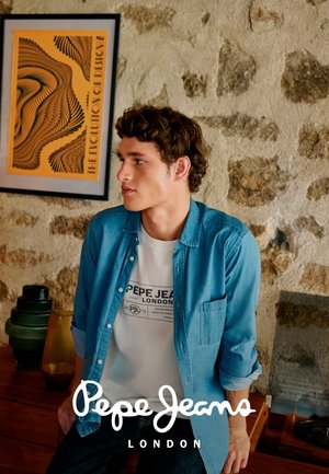 Junger Mann in weißem Pepe Jeans T-Shirt und blauer Jeansjacke sitzt an einer rustikalen Steinmauer, neben ihm abstrakte Kunst und Vasen.