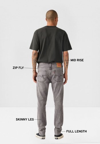 Hombre con tatuajes que lleva jeans ajustados grises de talle medio y longitud completa, y camiseta negra, etiquetados con las características: cremallera, talle medio, pierna ajustada, longitud completa.