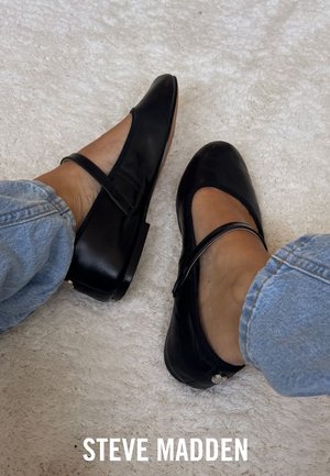 Zwarte leren ballerina's met een ronde neus, een bandje over de wreef en een lage hak, gecombineerd met lichtblauwe jeans op een gestructureerde witte ondergrond.