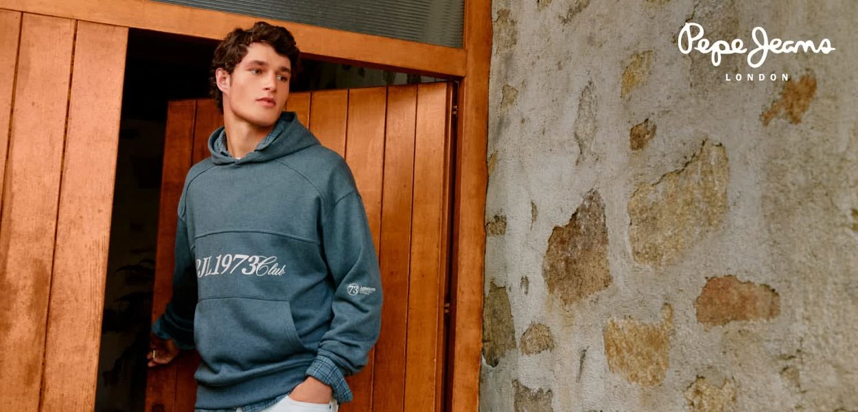 Sweat à capuche bleu avec impression frontale « PJ1973Club », doté d'une poche kangourou et d'une capuche à cordon de serrage, associé à un jean clair, devant une porte en bois.