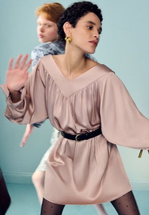 Zwei Frauen mit kontrastreichen Frisuren und Kleidung posieren dynamisch vor einem schlichten Hintergrund, eine in einem beigen Kleid, die andere in einem gemusterten Outfit.