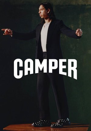 Person mit langen dunklen Haaren, schwarzem Anzug und weißem Hemd steht mit ausgestreckten Armen auf einem Holztisch und trägt schwarze Schuhe mit weißen Punkten.