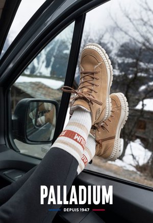 Beige samtige Knöchelstiefel mit weißen Gummisohlen, Schnürsenkeln und Reißverschlussdetails. Ein Paar gestreifte Socken mit roten und braunen Streifen, getragen mit schwarzen Leggings.